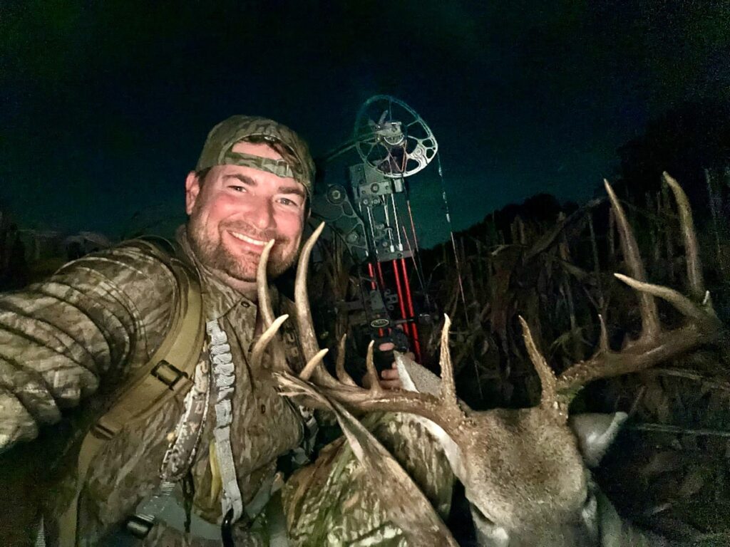 Bow hunter with harvested buck, large antlers visible, posing at night.