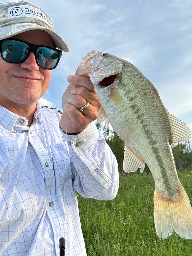 Angler holding a largemouth bass, wearing a Bois d'Arc cap and sunglasses.