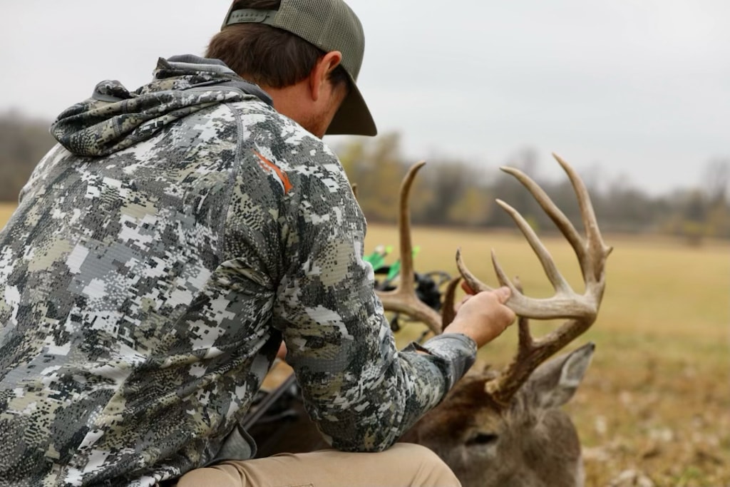 Hunter kneeling with a harvested buck, admiring its antlers. Camouflage hunting gear visible.