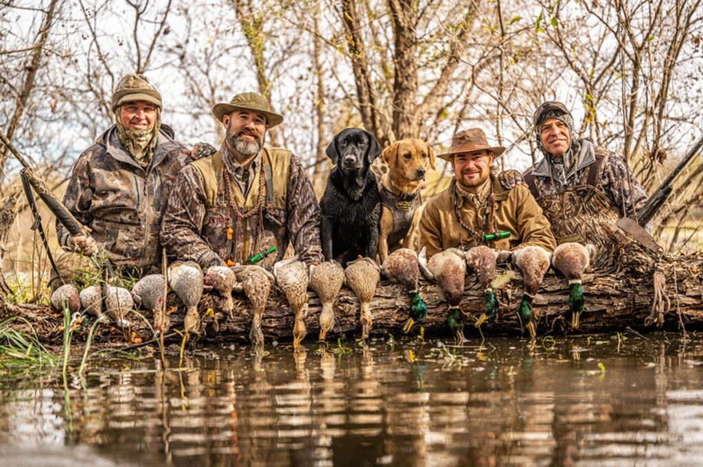 Hunters posing with hunting dogs and harvested ducks by a lake in autumn forest. Outdoor adventure, wildlife photography, duck hunting success.