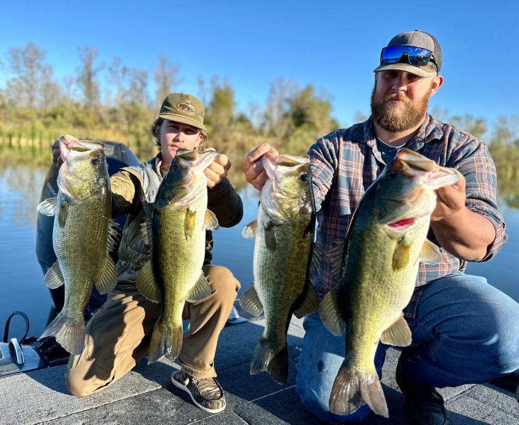 Two anglers proudly display large bass caught on a fishing trip, with clear blue skies and calm water in the background.