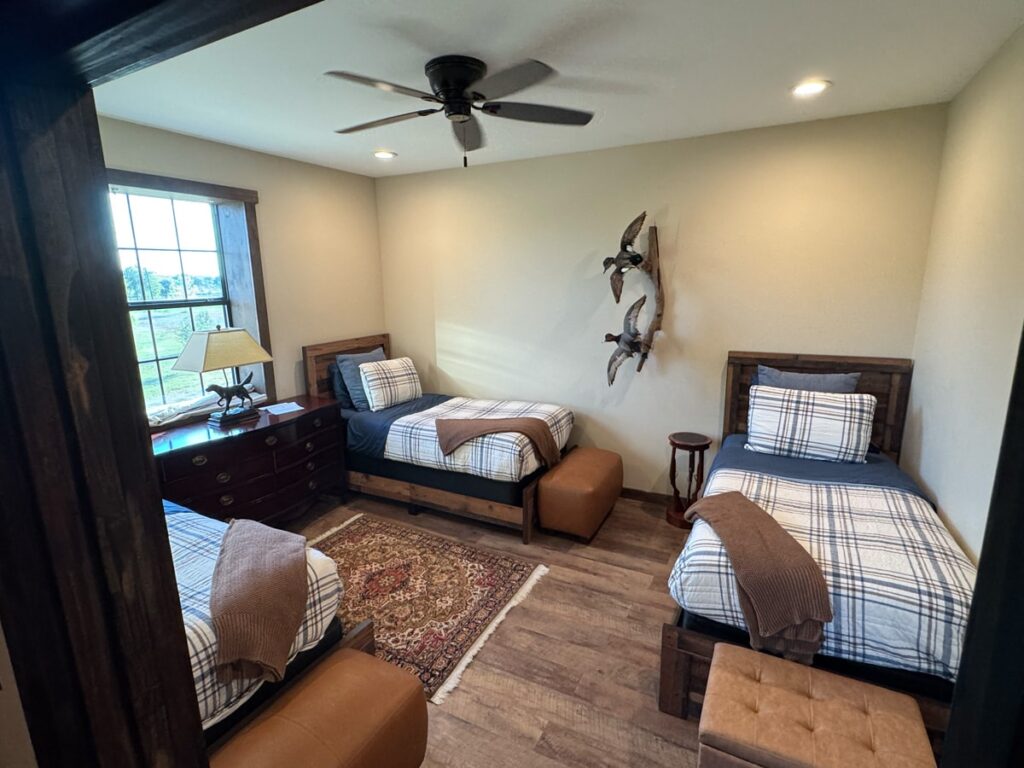 Cozy twin bedroom with wood accents, plaid bedding, and decorative wall ducks. Includes a window view, dresser, and ottoman for a rustic feel.