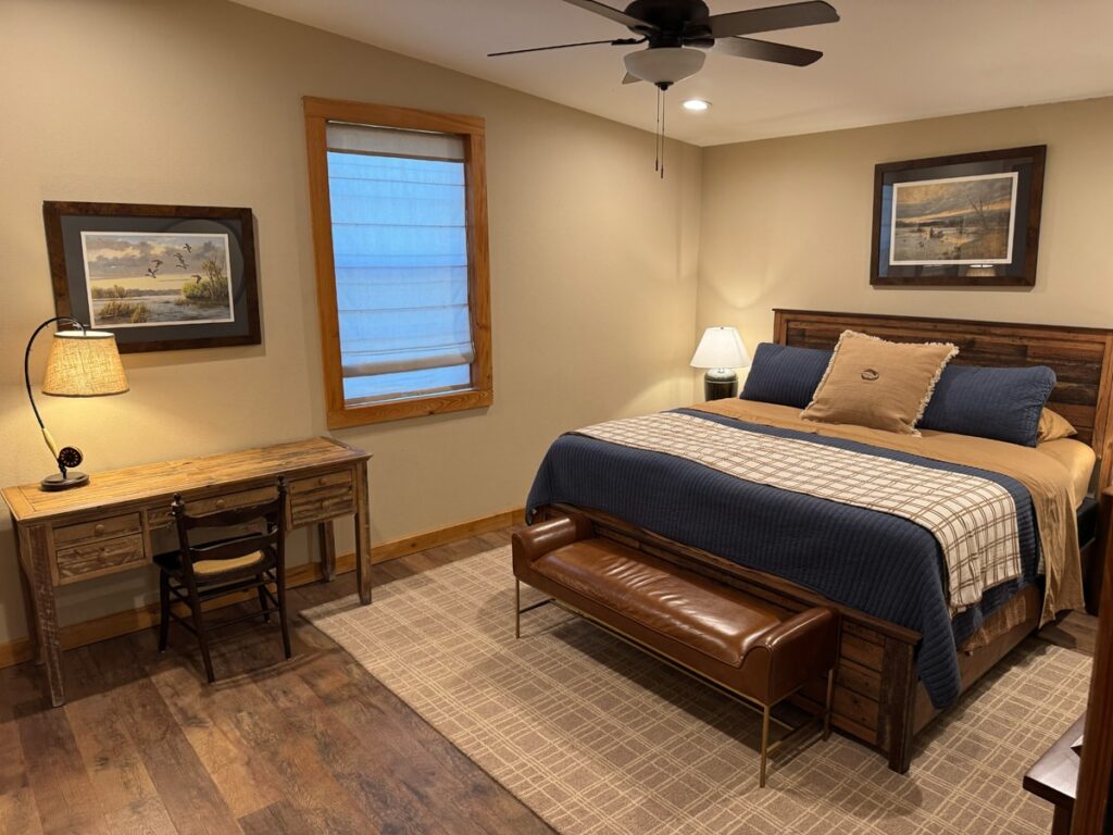 Cozy bedroom with a wooden desk and chair, framed artwork, and a neatly made bed with blue and beige bedding. Warm lighting creates an inviting atmosphere.