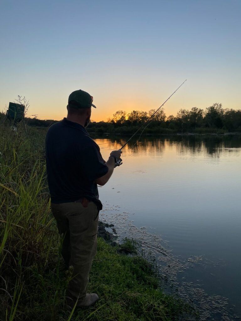 Man fishing at sunset, casting a line into a calm river surrounded by trees and tall grass.